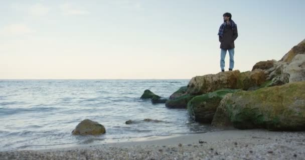 Schöner junger Mann, der auf den Felsen am Meeresufer steht. Bewölkter Himmel und kaltes Wetter. Draußen. Auszoomen — Stockvideo