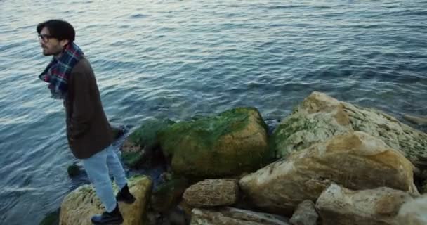 Blick von oben auf einen sympathischen Mann, der auf den Felsen am Meer steht und dem Meer gegenüber blickt. Kaltes Wetter. im Freien. — Stockvideo