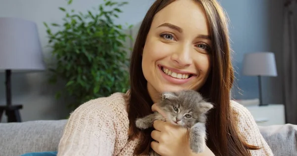 Retrato de una hermosa mujer joven abrazando a un gatito lindo y posando divertido a la cámara en la acogedora habitación en casa. De interior — Foto de Stock