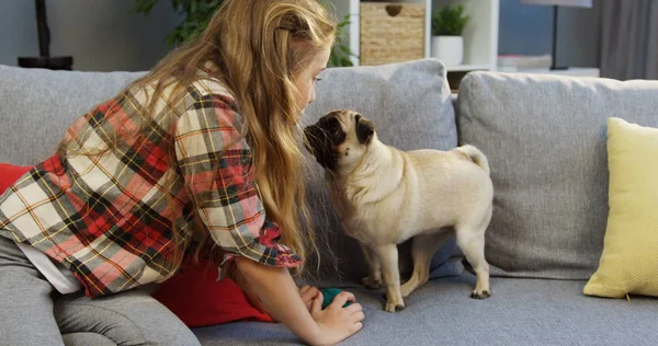 Chica rubia con un pequeño y agradable pug sentado en el sofá y mirándose en casa. En interiores — Foto de Stock