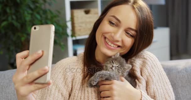 魅力的な若い女性がスマート フォンにかわいいキティ猫と素敵な selfies を作るします。肖像画を撮影します。居心地の良いリビング ルーム内 — ストック動画