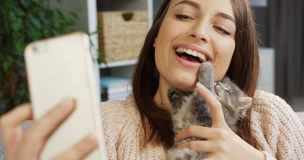 Vacker ung flicka gör selfies med en söt kattunge på den smarta telefonen medan du kitty spelar nära hennes ansikte. Närbild. Porträtt. Inne vardagsrummet — Stockvideo