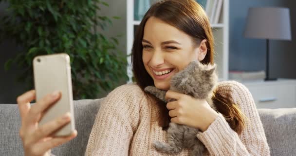 스마트 폰에는 귀여운 작은 고양이 함께 재미 있는 selfies를 만드는 매력적인 젊은 여자. 초상화입니다. 집에. 실내 — 비디오