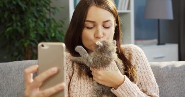 Привлекательная молодая женщина делает милые селфи с довольно маленьким котенком на смартфоне. Портрет. Гостиная. В помещении — стоковое видео