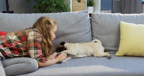 Маленька блондинка з милим мопсом сидить на дивані і погладжує його вдома. Внутрішні — стокове відео