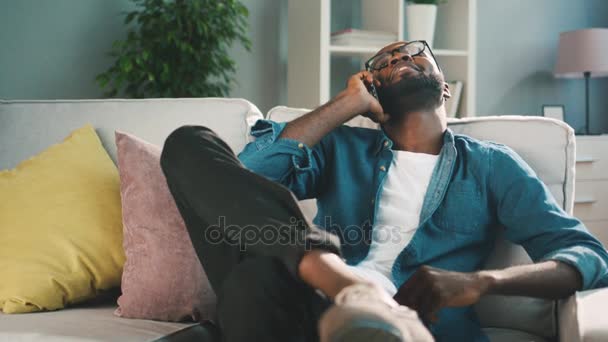 Afrikai fiatalember beszél barátommal szúró telefon. Szép fekete ember ül a kanapén, otthon és használ sejt telefon beszélő és barátja. — Stock videók