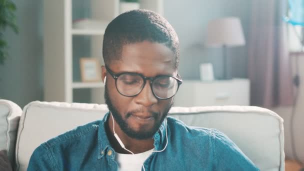Νεαρή μαύρη αφρικανική άνθρωπος, να ακούτε μουσική στα ακουστικά ενώ κάθεται στον καναπέ στο σαλόνι. Μαύρο άνδρα χαμογελώντας και χορεύοντας στο ρυθμό. Αργή κίνηση — Αρχείο Βίντεο