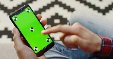 Erkek eller yeşil ekranlı siyah akıllı telefonu dikey olarak tutuyor ve kaydırıyor, bantlıyor ve yakınlaştırıyor. Bulanık halı arka planı. Omzundan. Krom anahtar. İzleme hareketi