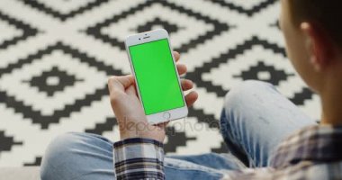 Beyaz adam elinde beyaz akıllı telefon yakın çekim. Bir dikey olarak tutan el ve başka bir kaydırma ve ekranda kaydediyor. Desenli halı arka plan bulanık. Omuz üzerinden