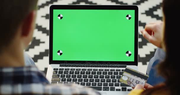 Uitzicht over de schouder op de paar zitten met een laptop voor hen en online met een credit card kopen. Groen scherm. Chromakey. Het bijhouden van beweging. Achtergrond met patroon tapijt — Stockvideo