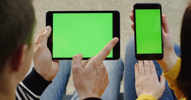 Weergave van de schouder van de mans handen scrollen op de zwarte tablet apparaat en dames handen op de zwarte smartphone met groen scherm. Chromakey. Close-up. — Stockvideo