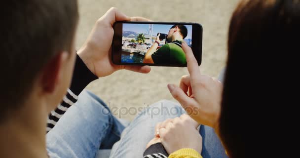 Нью-Йорк - Dec 01, 2017: Close up of the over shoulder view on the man and woman sitting hand in hand, scrolling photos on the black smartphone horizontally. Вид сверху — стоковое видео