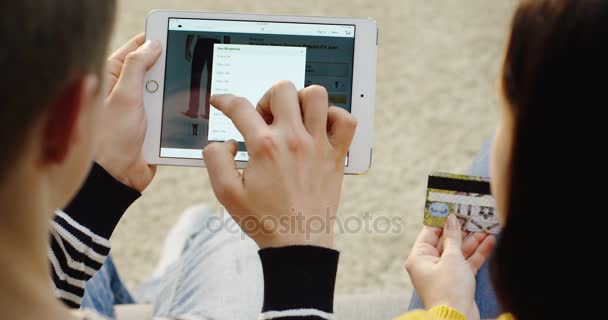 ニューヨーク - 2017 年 12 月 1 日: 上タブレット デバイスとクレジット カードで近くに座っている女性でアマゾンのウェブサイトでオンライン購入のための服のページをスクロール男に肩のビュー。クローズ アップ。ページのトップへ — ストック動画