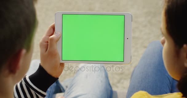 Вид через плечо на мужчину с белым планшетным устройством с зеленым экраном и женщиной, сидящей рядом с ним. Хрома-ключ. Закрыть — стоковое видео