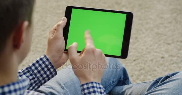 Uomo caucasico seduto con un dispositivo tablet nero e scorrimento e nastro su di esso. Sopra la spalla. Sfondo tappeto sfocato. Schermo verde. Chiave cromatica — Video Stock