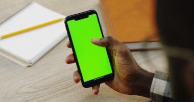 Afrikalı-Amerikalı dikey olarak yeşil ekran ile siyah bir akıllı telefon tutarak ve kaydırma eller mans. Chroma anahtar. Ahşap masa Notebook arka plan ile. Yakın çekim