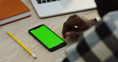 Beyaz ahşap masa ve Afrika kökenli Amerikalı adam üzerinde taping yatan siyah akıllı telefon yakın çekim. Omuz üzerinden. Yeşil ekran. Chroma anahtar.