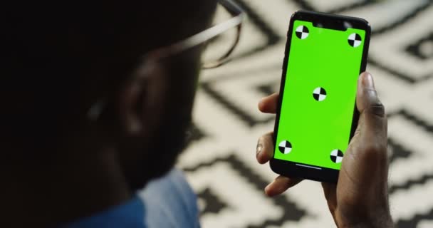 Sobre vista ombro no homem americano africano segurando um telefone inteligente preto verticalmente com tela verde e movimento de rastreamento. Chave Chroma. Fundo modelado — Vídeo de Stock
