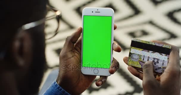 Vue sur l'épaule sur l'homme afro-américain tenant la carte de crédit et smartphone blanc avec écran vert verticalement et l'enregistrement sur elle tout en faisant des achats en ligne. Fond de plancher à motifs flous — Video