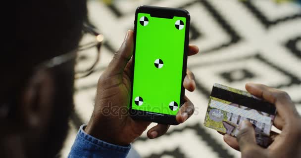 Vue de dessus l'épaule sur l'homme afro-américain tenant la carte de crédit et smartphone noir avec écran vert et le mouvement de suivi verticalement et défiler et enregistrer sur elle tout en faisant des achats en ligne. Flou — Video