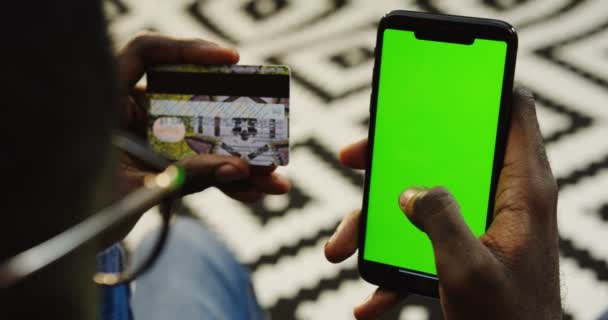 超过肩的看法, 非洲裔美国人的眼镜持有信用卡, 并输入其数据在一个黑色的智能手机, 在网上购物的垂直绿色屏幕。模糊花纹地毯 — 图库视频影像