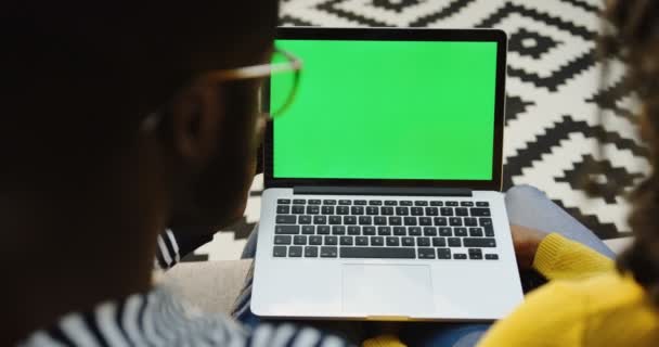 Sobre a vista do ombro do casal afro-americano assistindo algo no computador portátil com tela verde na frente deles. Chave Chroma. Piso modelado — Vídeo de Stock