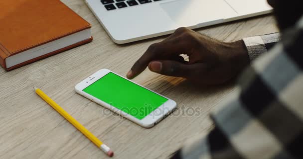 Primer plano del teléfono inteligente blanco que se encuentra en el escritorio de madera blanca cerca de cosas de oficina y afroamericano mano masculina grabando en él. Sobre el hombro. Pantalla verde. Clave de croma . — Vídeo de stock