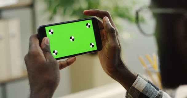 Πέρα από την άποψη ώμο στην αφρικανική αμερικανική αρσενικό χέρια κρατώντας μαύρα smartphone οριζόντια και δέσει σε αυτό στο δωμάτιο θολή φόντο. κοντινό πλάνο. Πράσινη οθόνη. Κλειδί Chroma. Παρακολούθηση κίνησης — Αρχείο Βίντεο