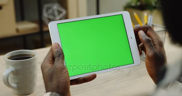 Sobre a vista do ombro no computador tablet branco segurado pelo homem afro-americano horizontalmente. Dedos rolando e gravando nele. Tela verde. Chave Chroma. Mesa de escritório de madeira com xícara de café — Vídeo de Stock