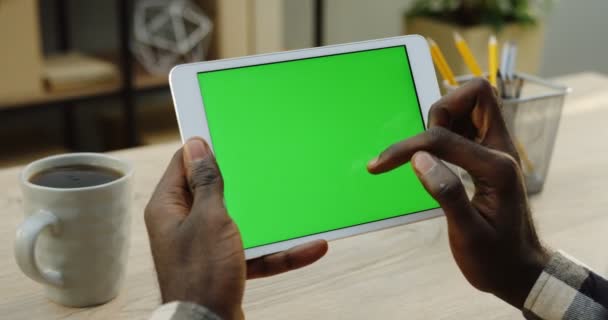 Computador tablet branco em mãos de homens americanos africanos horizontalmente. Dedos rolando e gravando nele. Tela verde. Chave Chroma. Mesa de escritório de madeira com xícara de fundo de café. Fechar — Vídeo de Stock
