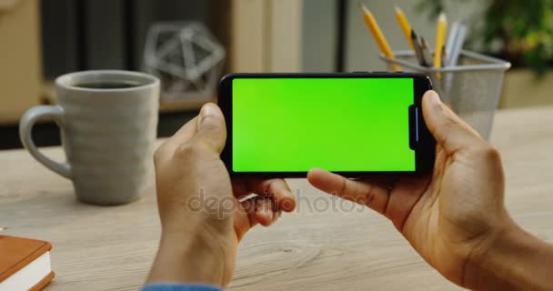 Το Top view για την καυκάσια επανδρώνει τα χέρια κρατώντας οριζόντια μαύρη smartphone το ξύλινο γραφείο με φλιτζάνι καφέ φόντο. Κοντινό πλάνο. Πράσινη οθόνη. Κλειδί Chroma. — Αρχείο Βίντεο