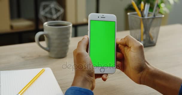 Smartphone blanco siendo utilizado por manos masculinas verticalmente. Los dedos se desplazan y pegan en él. Pantalla verde. Clave de croma. Mesa de oficina de madera con taza de fondo de café. De cerca. — Vídeo de stock