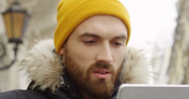 Tablet bilgisayarda kış gününde kaydırma sarı şapkalı sakallı genç beyaz adamın kapatın. açık havada. Portre