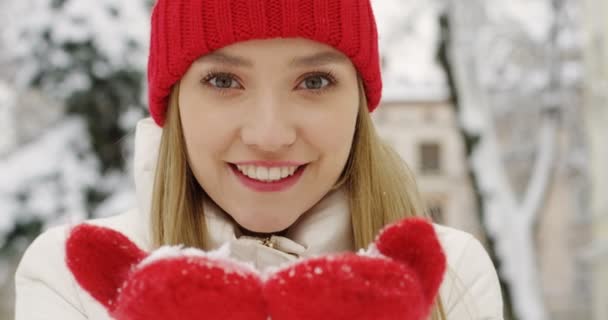 赤い帽子と手袋カメラに手の手のひらで 雪と冬の日の町の通りにそれを吹いて魅力的な金髪女性のクローズ アップ 肖像画 — ストック動画