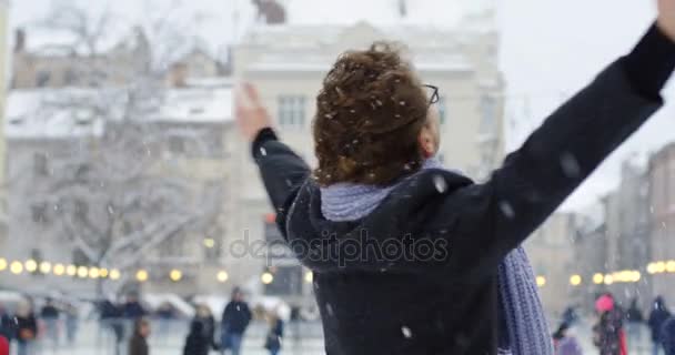 カジュアルなスタイルを手で転がしながら ヨーロッパの町の広場に雪が降っている冬の間にガラスの幸せな魅力的な男 — ストック動画