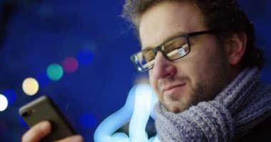 Çekici adamın gözlük ve eşarp hilding siyah smartphone ve üzerine bantla kapatın. Bulanık Noel ışıkları ile geç akşam. portre. Dışarıda