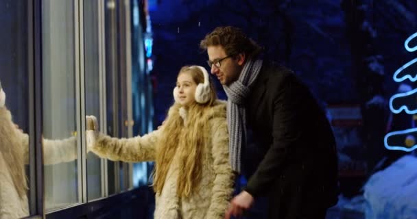 年轻可爱的女孩与长的金发碧眼通过窗口的情况下 与她的父亲在傍晚的圣诞装饰街的东西 — 图库视频影像