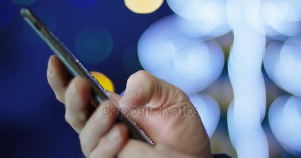 Close Male Hands Scrolling Black Smartphone Blurred Christmas Lights Background — Stock Video