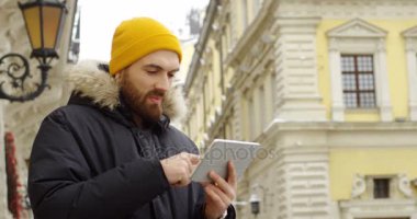 Sarı şapka ve kaydırma ve yakınlaştırma onun tablet cihaz ekranında soğuk kış gününde kasabada sıcak kış ceket sakallı genç çekici adam. Açık. Portre vurdu