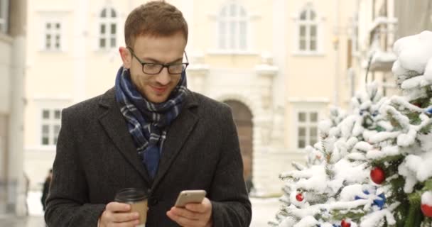 メガネ手にコーヒーを飲みながらの冬の街でうれしそうに歩いて 彼のスマート フォンにテーピングで魅力的な男性 背景にクリスマス ツリー — ストック動画