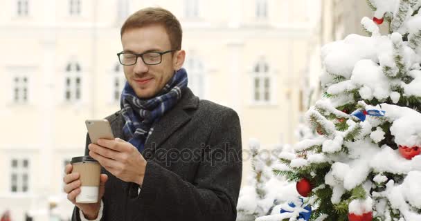 肖像拍摄的迷人的白种人在眼镜微笑着 站在附近的圣诞树与咖啡走在一个手和手机在另一个 镇街道 — 图库视频影像