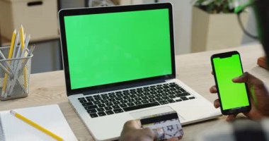 Omuz üzerinden Afrika kökenli Amerikalı görünümü kaydırma ve o süre alışveriş online kredi kartı tutarak iken yeşil ekran siyah bir smartphone üzerinde taping eller mans. Olan bir dizüstü bilgisayar