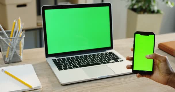 クローズ アップ スクロールと緑色の画面で黒のスマート フォンとラップトップ コンピューターをオフィスのテーブルの上の緑色の画面でテーピングが背景に手マンします クロマ — ストック動画
