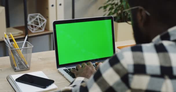 アフリカ系アメリカ人の肩のビューの上の作業と緑色の画面の横に横になっている白いスマート フォンとラップトップ コンピューターのキーボードでテーピングを男します クロマ 近代的なオフィスの背景 — ストック動画