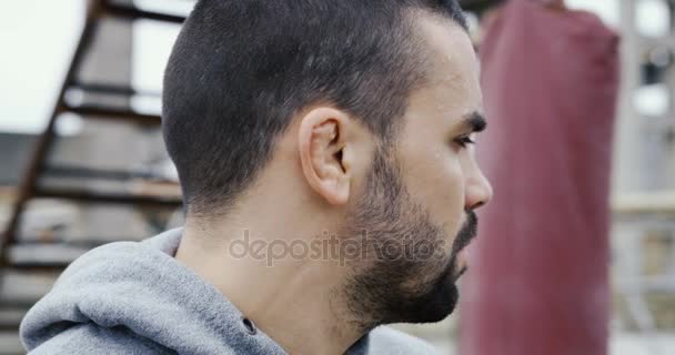 Sportsmans 的脸上有一张黑色的胡须 一边看着 一面严肃地看着镜头 拳击袋和废墟上的模糊背景 冷的灰色天气 — 图库视频影像