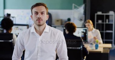 Beyaz tişörtlü kamera ve bulanık office arkasındaki bilgisayarlar işçilerle bakarak çekici genç adam portresi. Kapalı
