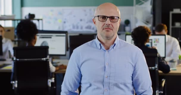 Retrato de un hombre calvo y guapo con gafas mirando con una sonrisa a la cámara y cruzando sus manos La oficina borrosa con los trabajadores en el fondo de las computadoras. En interiores — Vídeo de stock