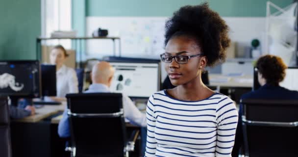 Retrato de una joven afroamericana atractiva con gafas mirando al costado y que sonriendo a la cámara. La oficina borrosa con los trabajadores en el fondo de las computadoras. De interior — Vídeo de stock