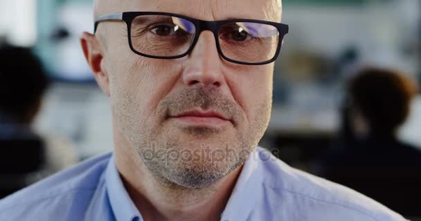 Primer plano del hombre caucásico de mediana edad en gafas mirando a un lado y que sonriendo a la cámara. La oficina borrosa con los trabajadores de fondo. Retrato — Vídeo de stock