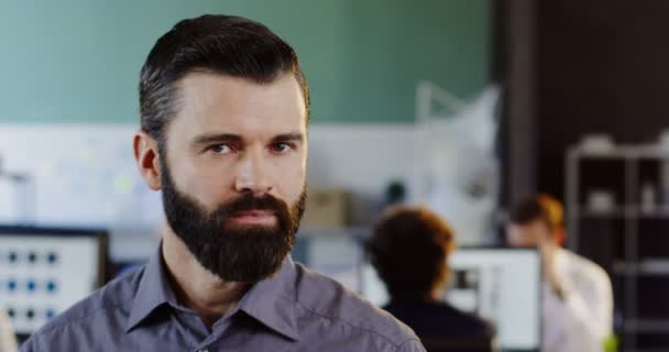 Primo piano di attraente giovane uomo con la barba guardando e sorridendo nella fotocamera. L'ufficio offuscato con persone con background informatico. Dentro. Ritratto — Video Stock